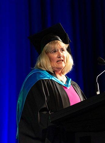 A blonde woman wearing academic regalia is standing at a podium